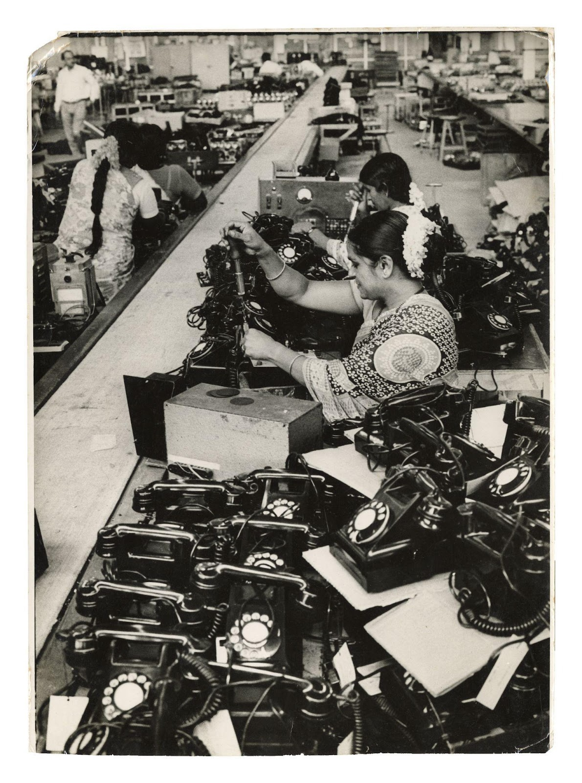 In this black and white photograph we see women working in a telephone factory. There are four women working in the photograph. They are dressed in patterned saris, simple jewellery and flowers in their hair. In the top-left corner, there’s a smaller figure of a formally dressed man walking past, suggesting routine surveillance by a senior. 