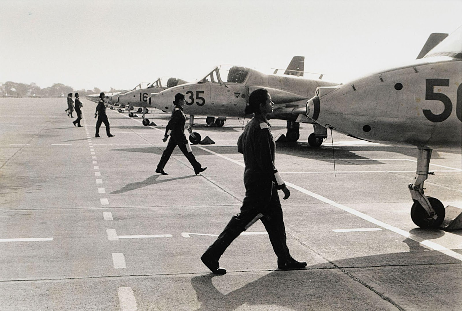 A black and white photograph of a  series of uniformed female pilots march towards their individual jets waiting on the tarmac.