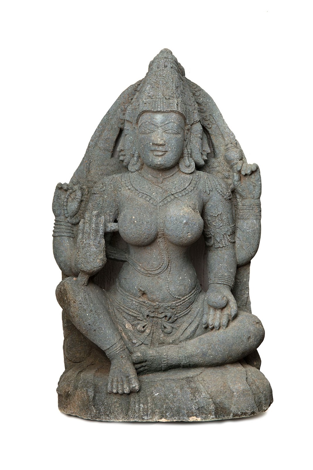 A grey steatite schist sculpture of a female with three faces and four hands. She is seated with her eyes closed and each of her three heads has a crown on it. 