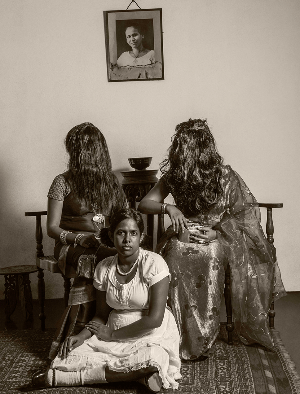 A woman in a dress sits cross legged on a carpet. Behind her are two saree-clad women seated on chairs, with their hair completely covering their faces. 
