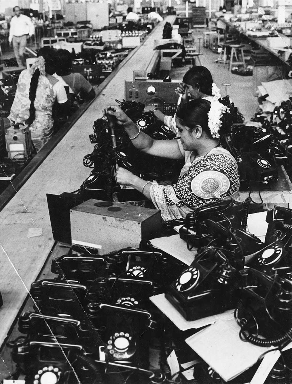 In the foreground are a number of old-school telephones with rotary dials. Behind, women with flowers in their hair sit next to each other as they assemble more such telephones. One man is seen in the background, at the top left corner.