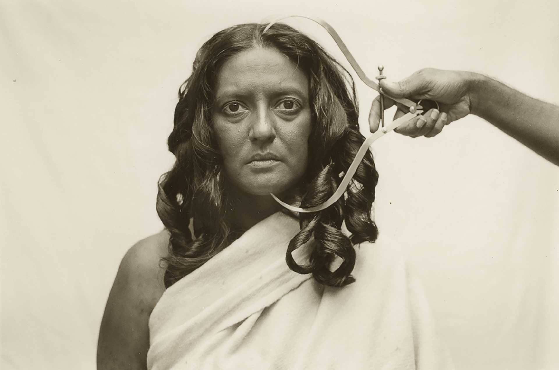 In the centre, a woman with hair curled in large ringlets looks straight ahead. From the right, a hand emerges holding a measuring device next to her face.