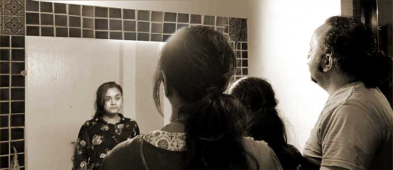 The back view of three people – a woman, a man and a girl standing between them – all looking into a mirror. The photograph is edited such that only the girl’s reflection is seen in the mirror.