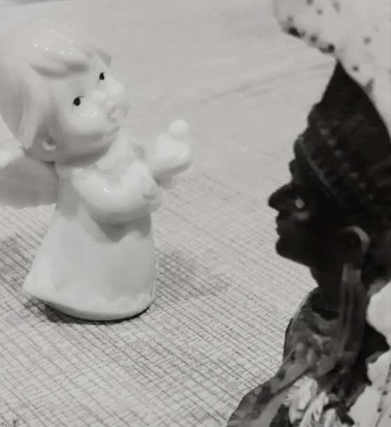 A black and white photograph in which a white porcelain doll, shaped like an angel, faces a black figurine with headdress and jewellery.