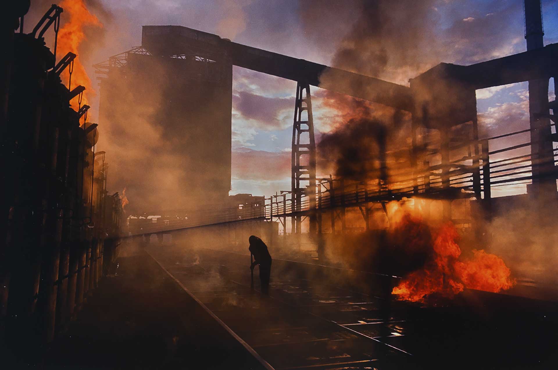 Silhouette of a person sweeping railway tracks, with large fires burning on either side of them. The fires are bright orange and produce thick black smoke. There are large industrial-like structures in the background.