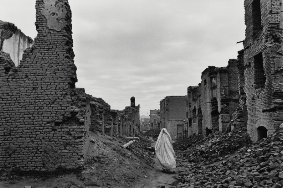 Rear view of a person walking in between the ruins of bricked buildings with rubble all around. They are wearing a white chador (traditional Afghani full body cloak).