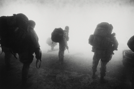 Three armed men in uniforms, carrying large bulky backpacks, are moving in a triangular formation. In the background distance, only a white haze and perhaps part of a fourth figure can be seen.