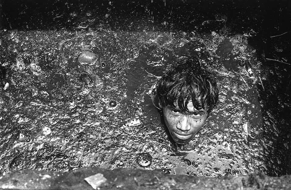 The head of a young man, as if floating, surrounded by murky sewage water. He seems to be looking up.