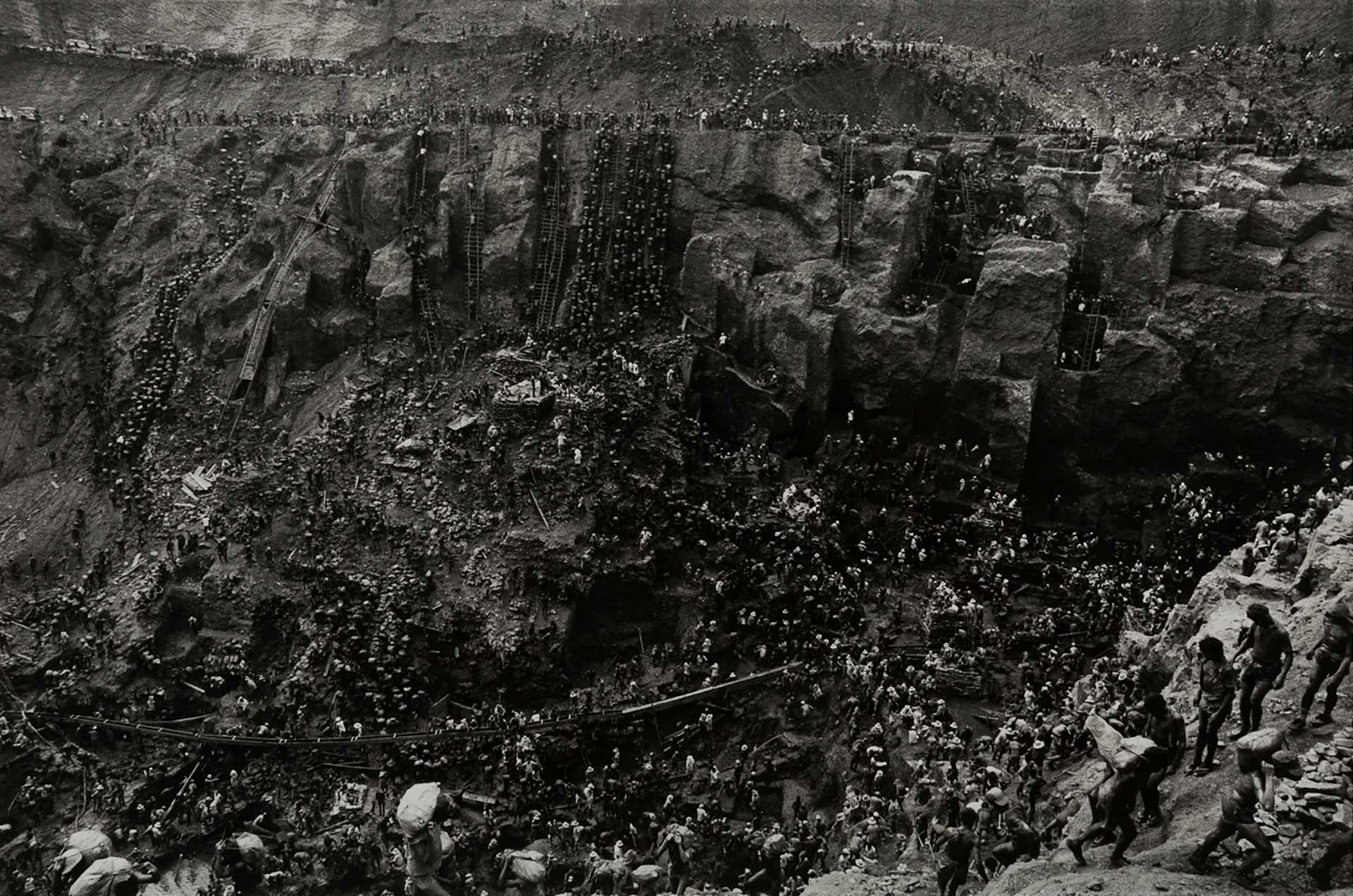 An aerial view of a huge crowd of people working in a mine. Makeshift ladders, bridges and people carrying heavy sacks are seen.