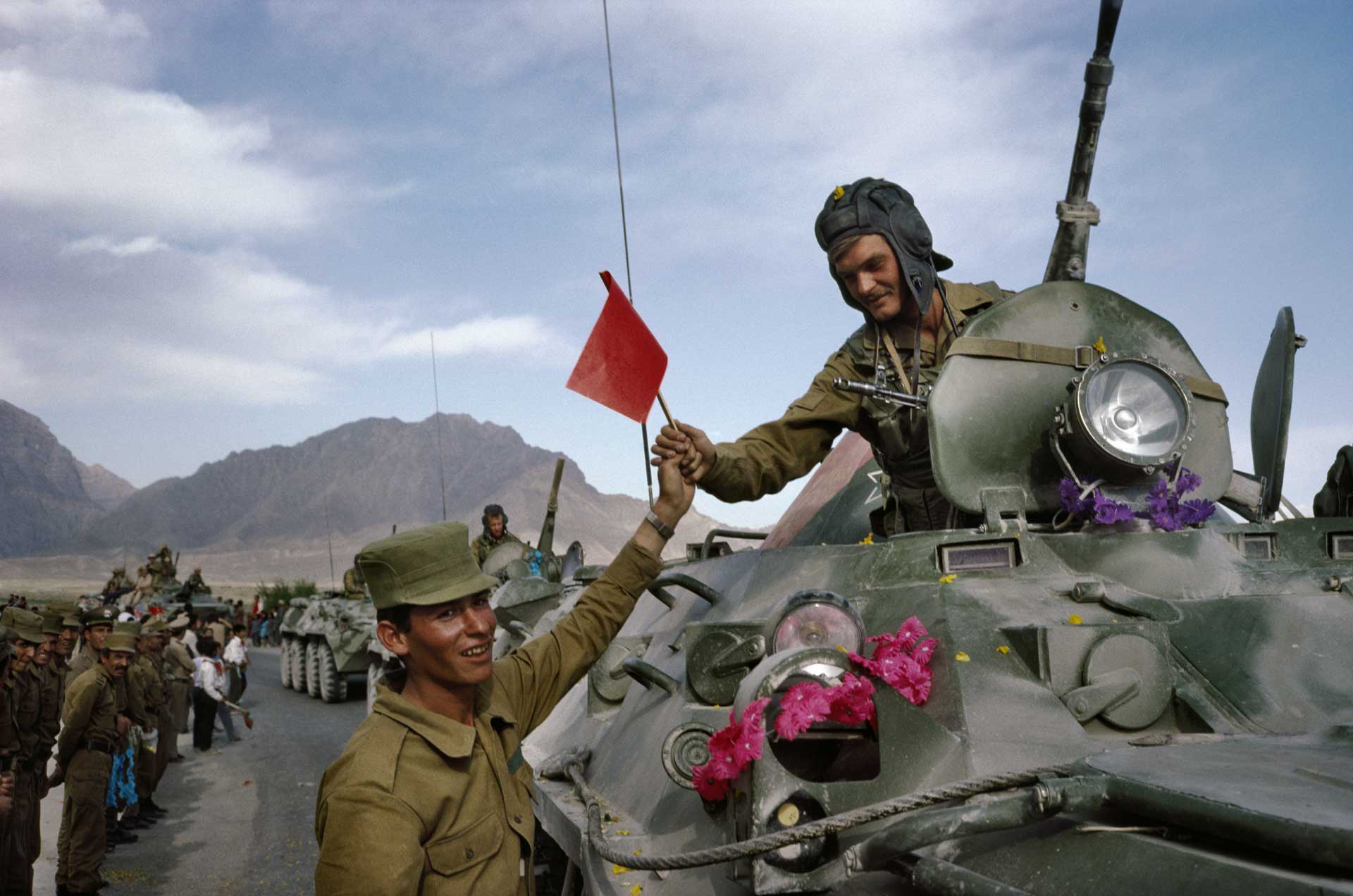 In the foreground, a soldier in a tank is handing over a red flag to a smiling soldier standing below. In the background, men in uniform stand by the side of the road, watching as the tanks pass by.