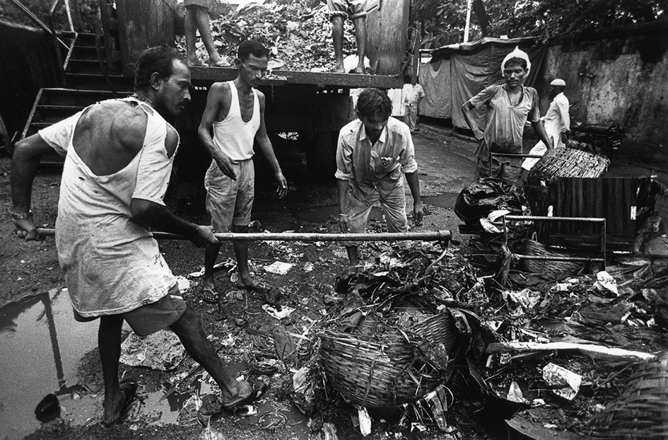 Four men stand around a pile of garbage. The man on the left is raking it while wearing a torn t-shirt, revealing part of his skeletal back. Another is leaning on a cart. There is a garbage truck in the background.