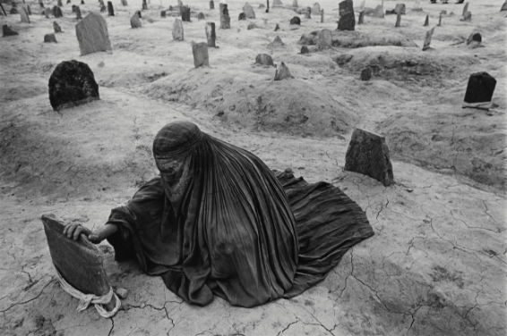 A woman wearing a black chador (traditional Afghani full body cloak) puts her hand on a gravestone as she sits on an uneven, cracked surface. Around her, the ground is littered with headstones marking many graves.