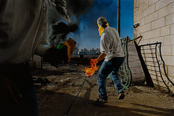A man wearing a cap and mask covering his face is seen from behind. His left hand looks aflame as he appears to be throwing an object on fire. In the foreground, on the left edge, is part of another person who also seems to be holding an object on fire that’s emitting thick grey smoke.