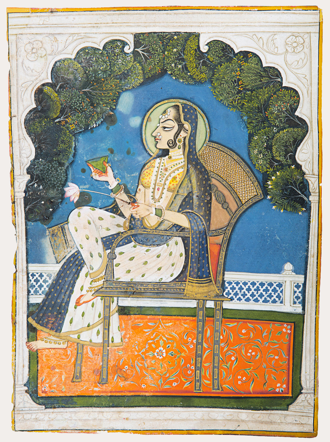 An outline of a window inside which a woman looks sideways and holds a betel leaf in one hand and a lotus stem in the other. She folds her leg over the other. Her background is an evening sky and trees. She is seated on a high chair on an orange carpet with floral design. (how do we say, we can see her from a window)