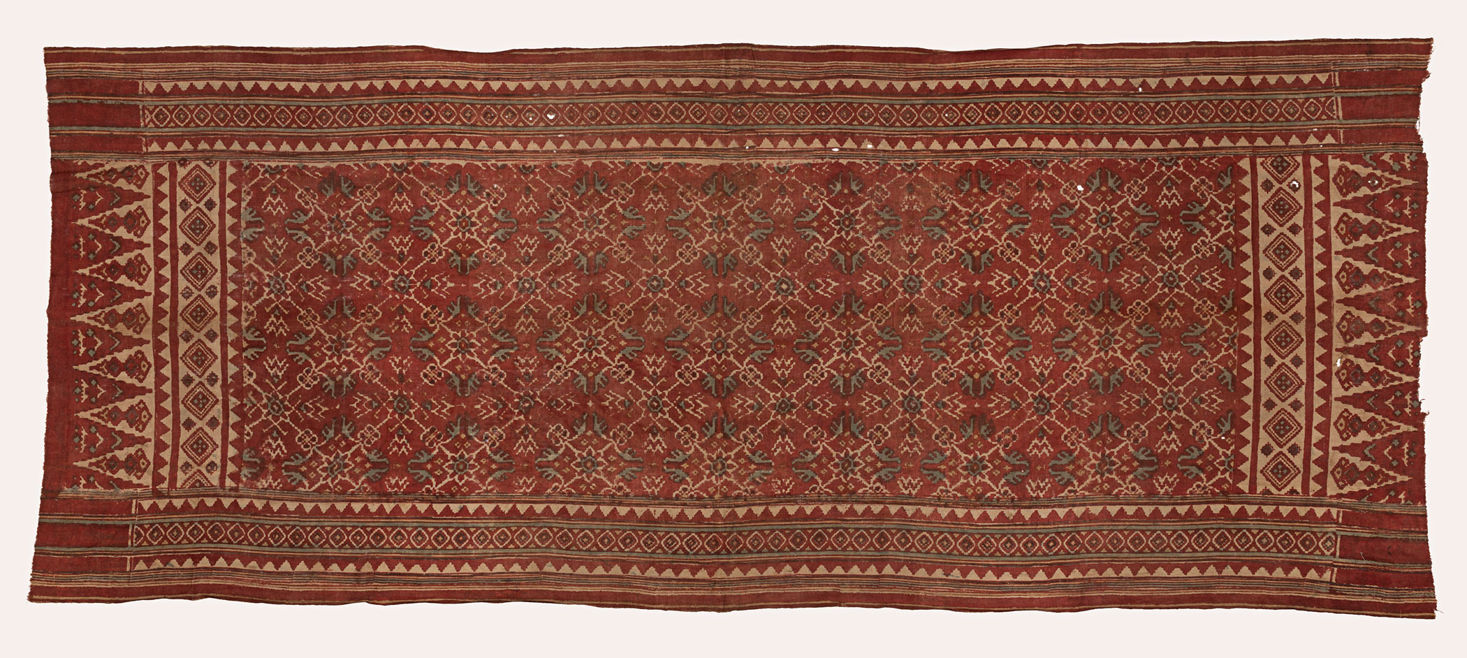 A rectangular cloth with red, green, off white faded patterns. The narrow ends have long triangular patterns while the wider borders have symmetrical triangles between horizontal bands of intricate motifs.
