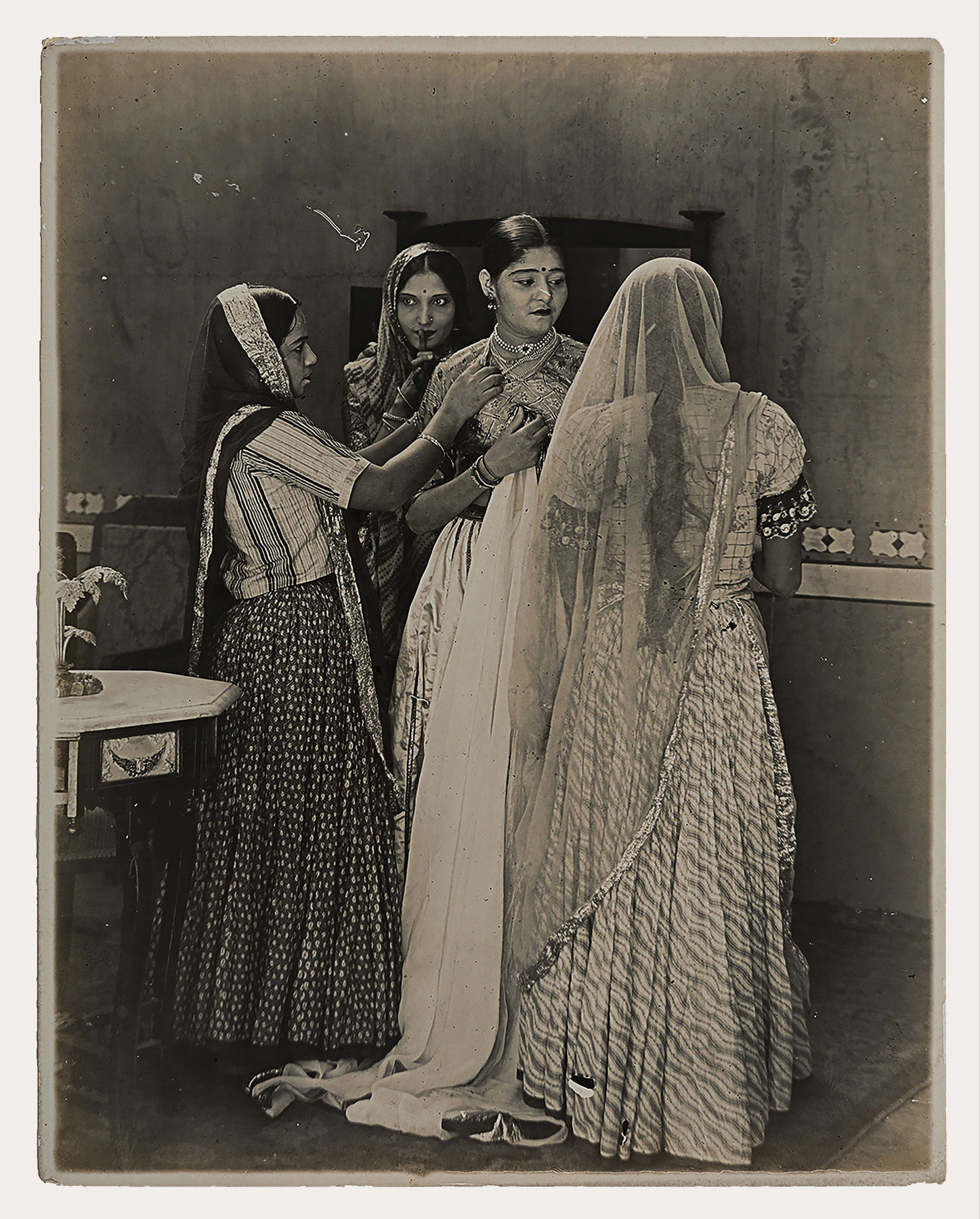 A woman stands in an enclosed space with a tilted head, holding a long piece of fabric close to her chest. In front of her, is the back of another woman and beside her, is a different woman trying to adjust her jewelry, facing sideways. Behind the three women is a fourth woman with her hand on her lips.