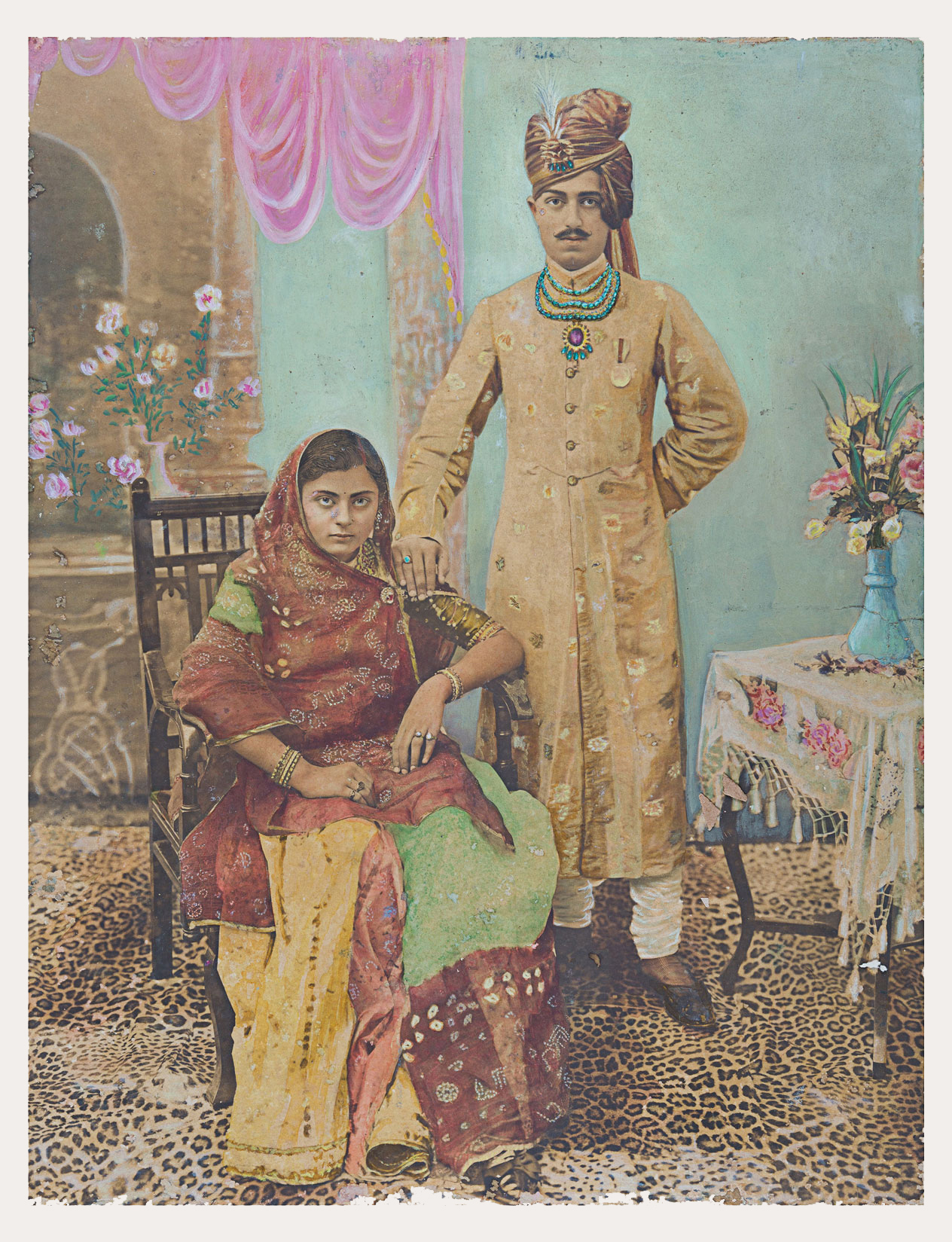 A woman looks directly at us while comfortably seated on a chair. A man wearing an intricate turban and jewelry is standing next to her, with his right hand on her shoulder. They are in an enclosed space which is upholstered with a carpet with leopard skin design.