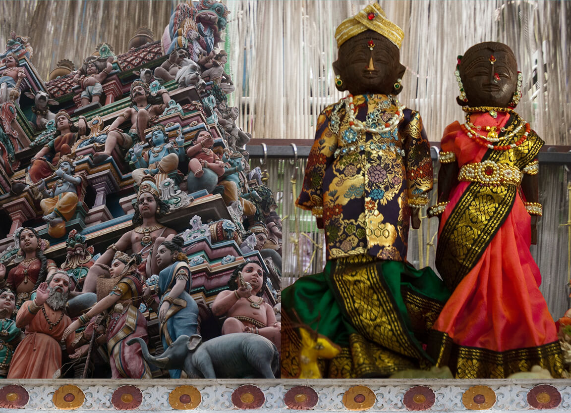 Two wooden dolls dressed in colourful clothing and jewelry . Next to it is the top of a south Indian entrance to a temple (gopuram).