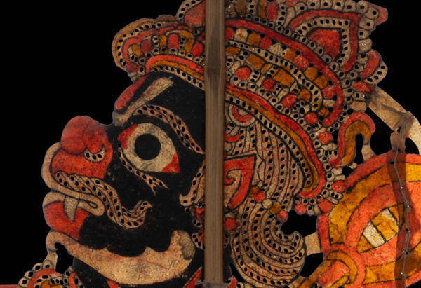 A detail of a puppet called Anjaneya looks towards the left with his eyes popping out. He is painted in yellow, black and orange paint and is attached to a wooden stick for the puppeteer to control his movements.