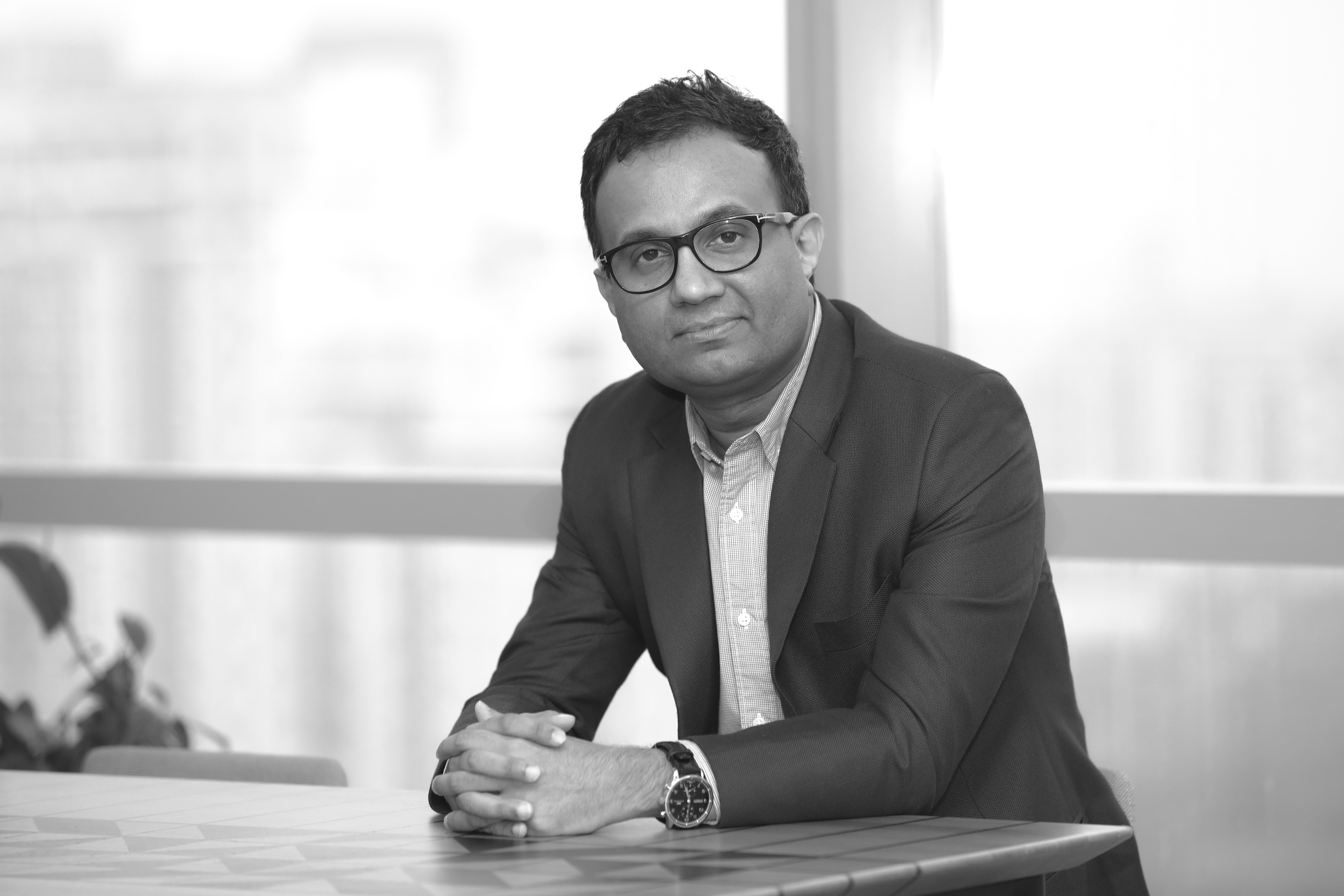 Ajit in black and white wearing a suit, he wears glasses and a watch on his wrist. He looks into the camera smiling slightly.