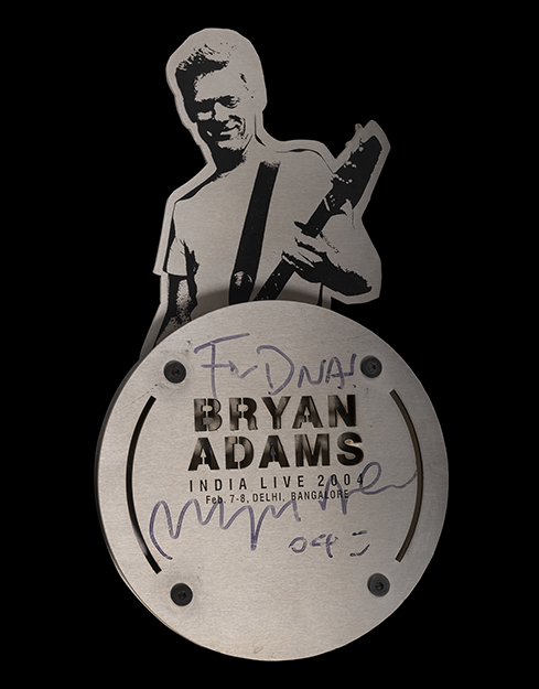 Illustrated black and white Bryan Adams holds a guitar with “for DNA, Bryan Adams, India Live 2004” written below it.