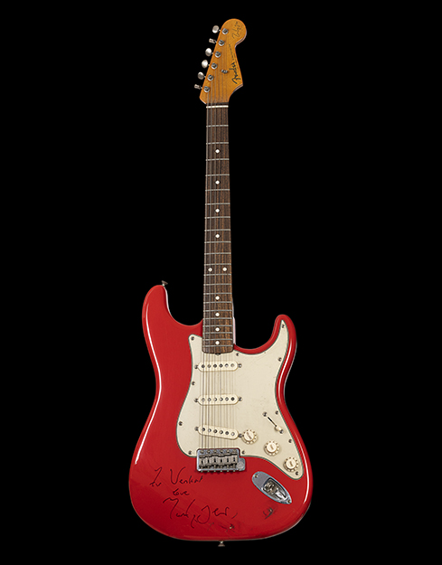 A bright red guitar with white design that takes the shape of the guitar. At the bottom of the guitar is a note which says “to Venkat, love Mark.”