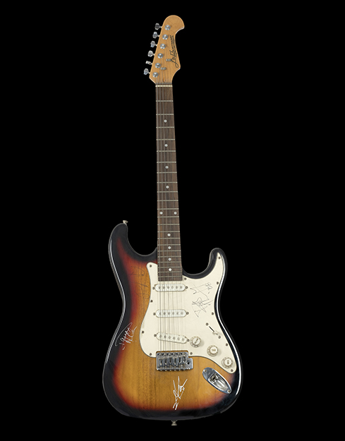 A guitar with a gradation of brown and black fading at the edges. Scribbled autographs scattered everywhere. The base of the guitar has a logo J&D by Jack and Daniel brothers.