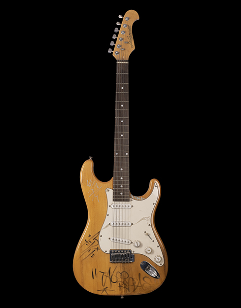 A brown and white guitar with a roundish base and prominent circular tone and volume regulators. The head of the guitar has the logo J & D, by Jack and Danny brothers.