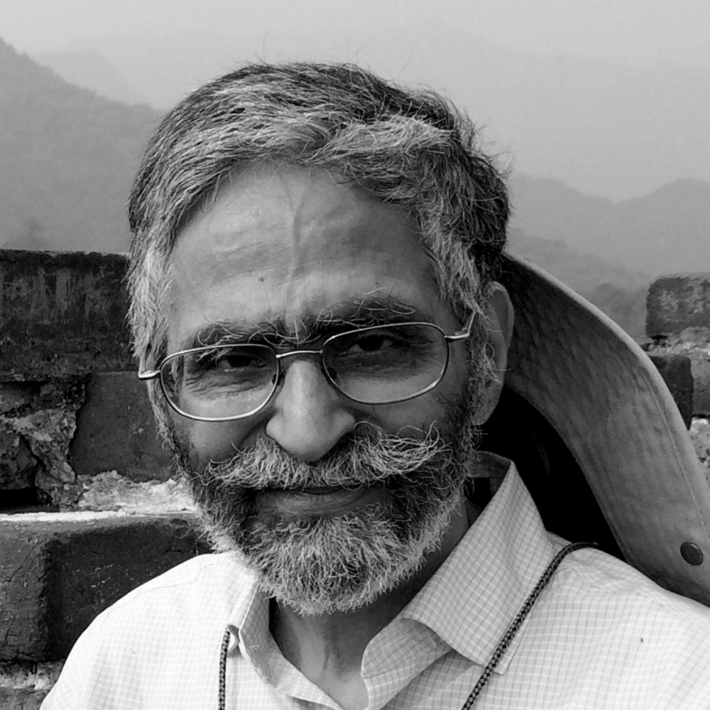 man with a slight smile His hair is greying he wears glasses. He has facial hair. Wearing a hat around the back of his neck and a white collared shirt