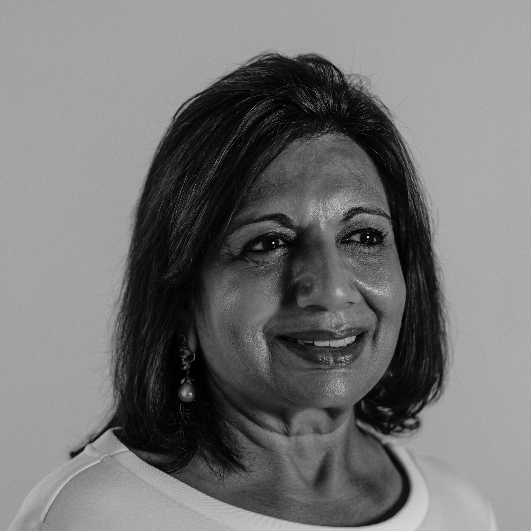Kiran Mazumdar Shaw in black and white smiling away from the camera, she has chin length hair and is wearing a white round neck shirt and drop earrings.