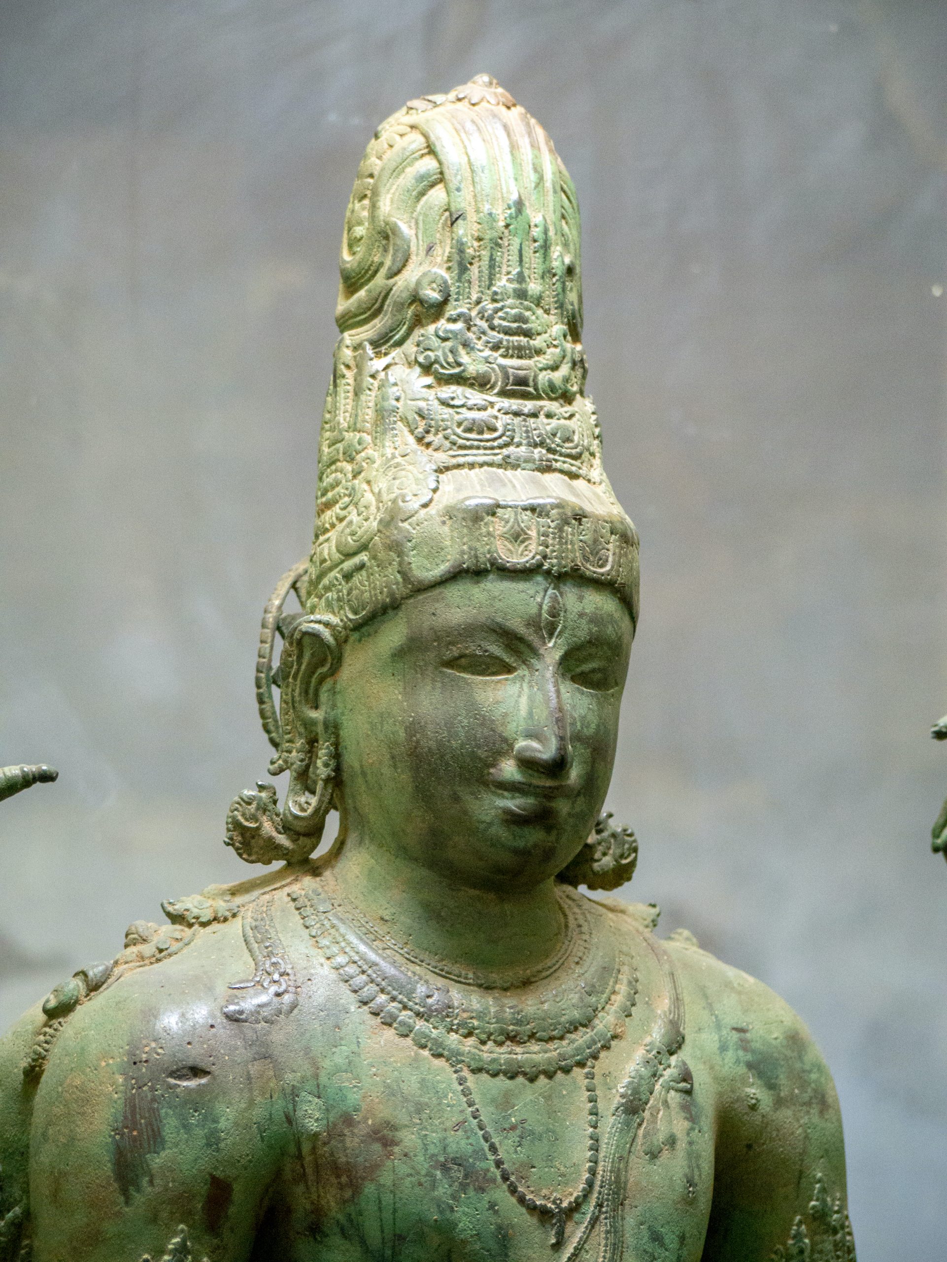 Bronze sculpture wearing several ornaments. Has oxidised to a green colour with patches of bronze seen on the head and chest of the sculpture.