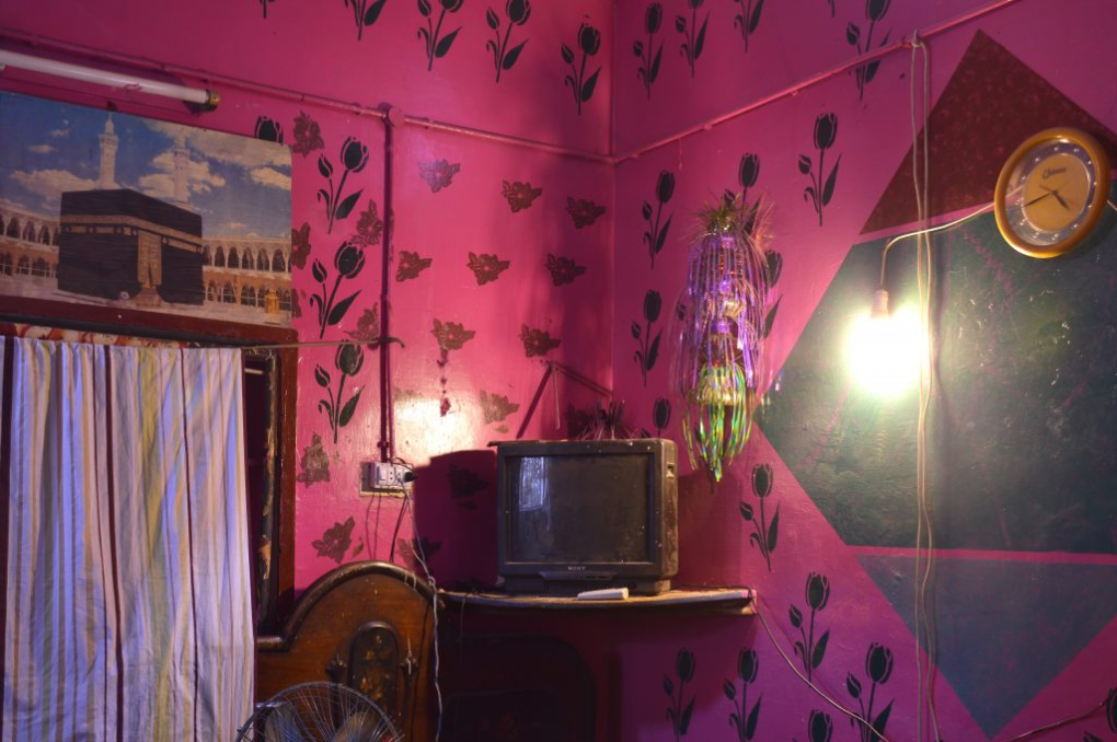 An old television in a pink walled room, black flowers painted on the wall and a clock on the right side. hanging lantern next to the tv.