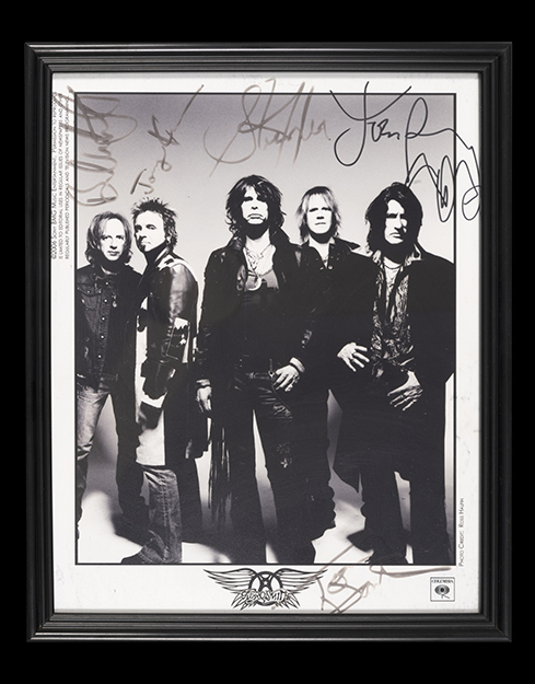 The band members of Aerosmith stand next to each other. Their silhouettes are dark grey against a faded light grey background with scattered autographs above them.