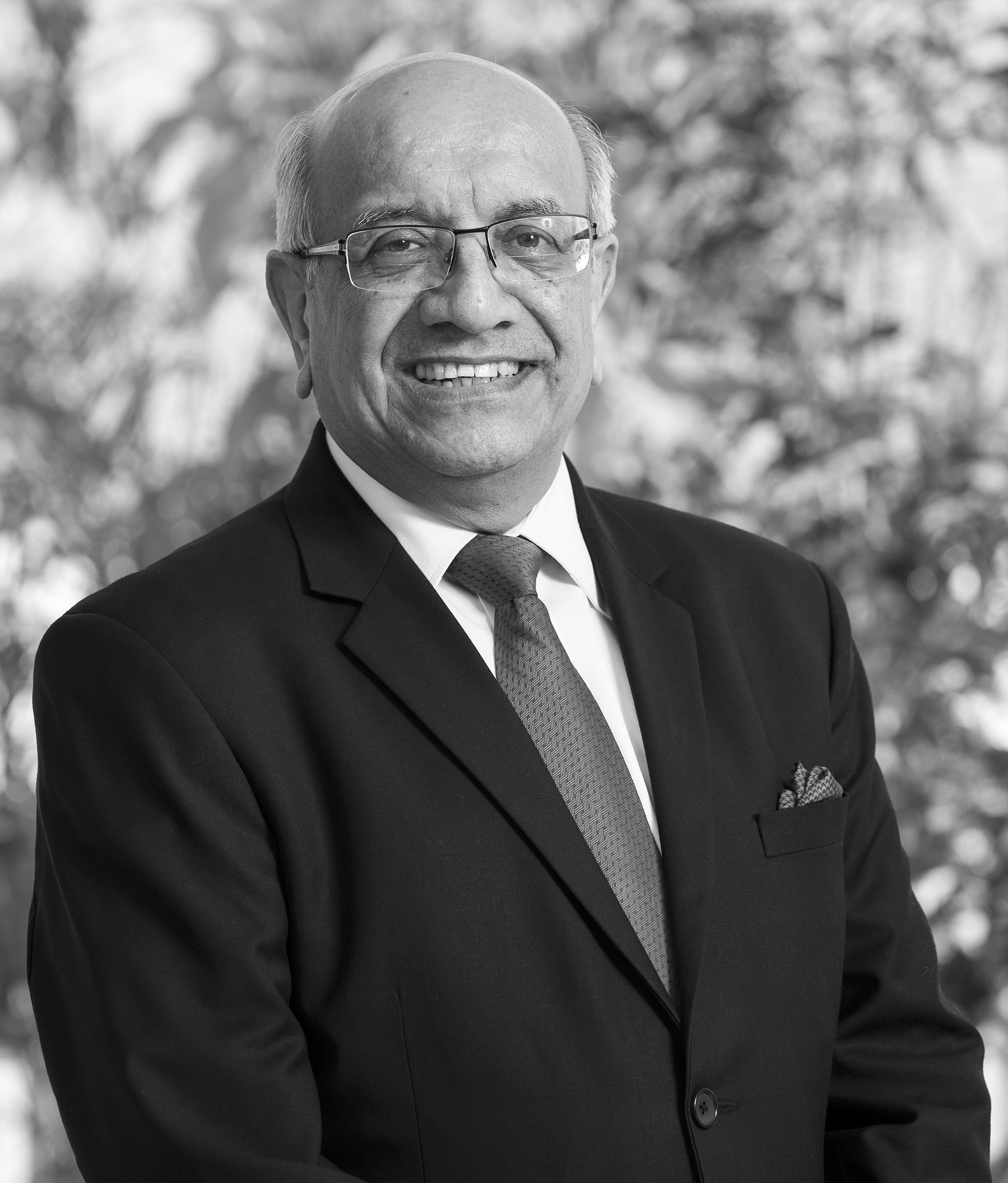 Som Mittal in black and white wearing a suit, tie and glasses, standing at an angle and looking into the camera smiling.