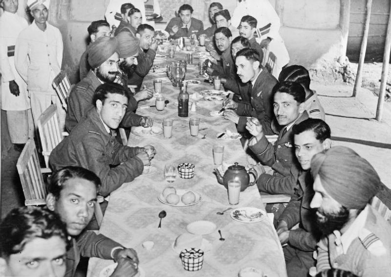 In Black and white, a group of army officers sitting around a long dinner table in an army mess, some looking directly into the camera