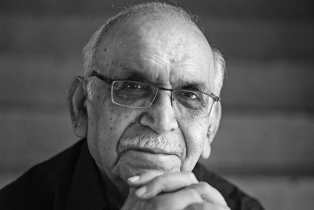 BN Goswamy in black and white with a moustache, wearing glasses, a black shirt and looking at the camera smiling with fingers joined under his chin.