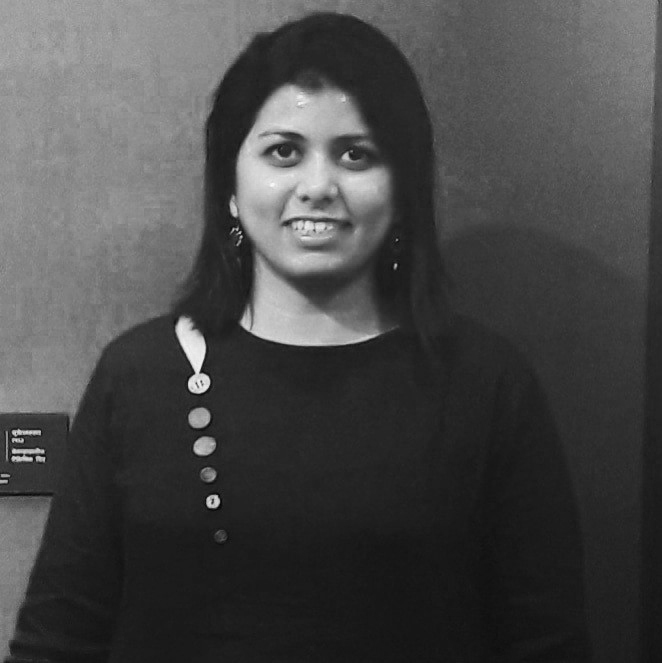 Madhura in black and white has straight hair until her shoulders is wearing a black blouse with button detailing. She is facing the camera and is smiling.
