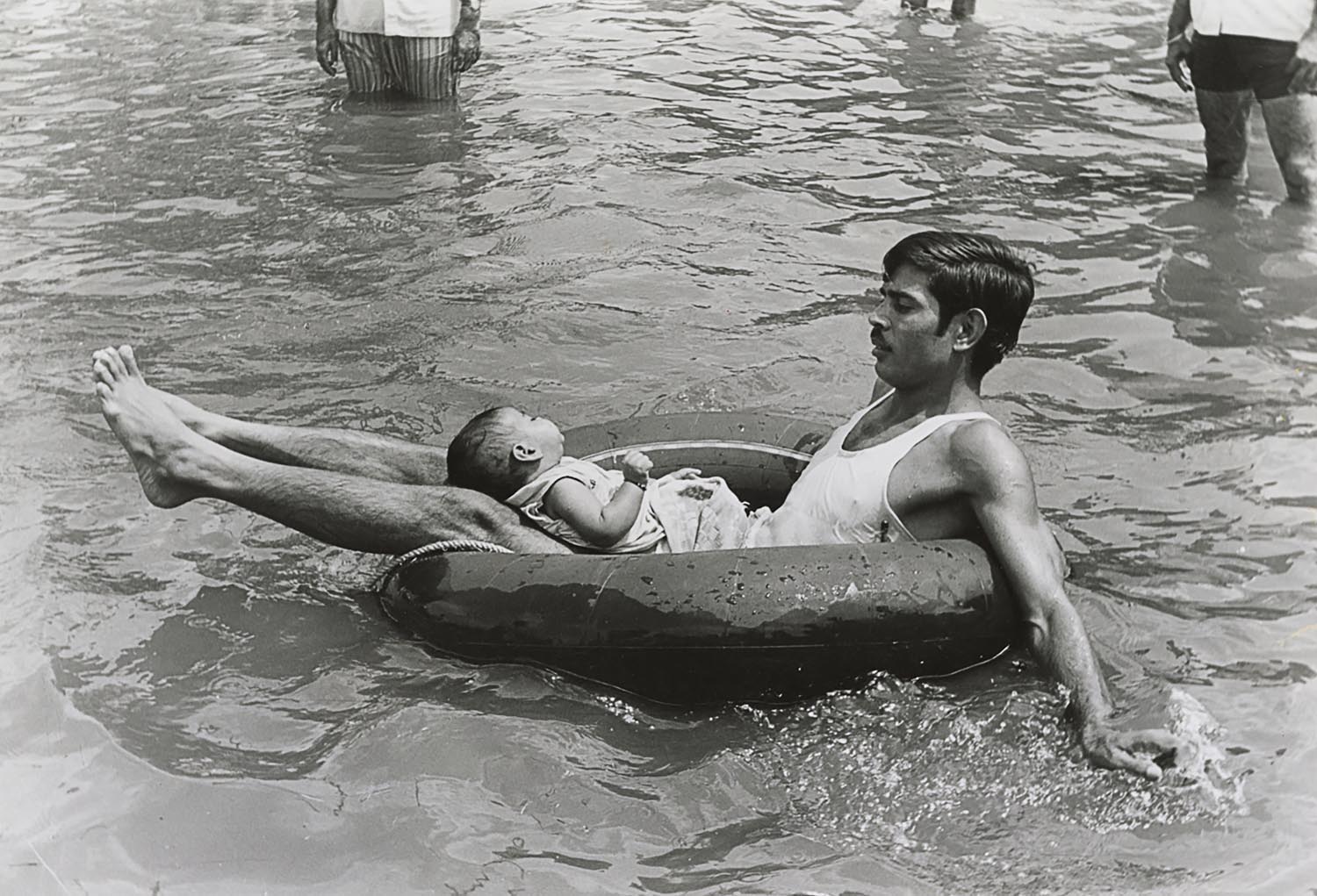A man on a rubber float with a baby on his lap, carrying themselves to safety during a flood.