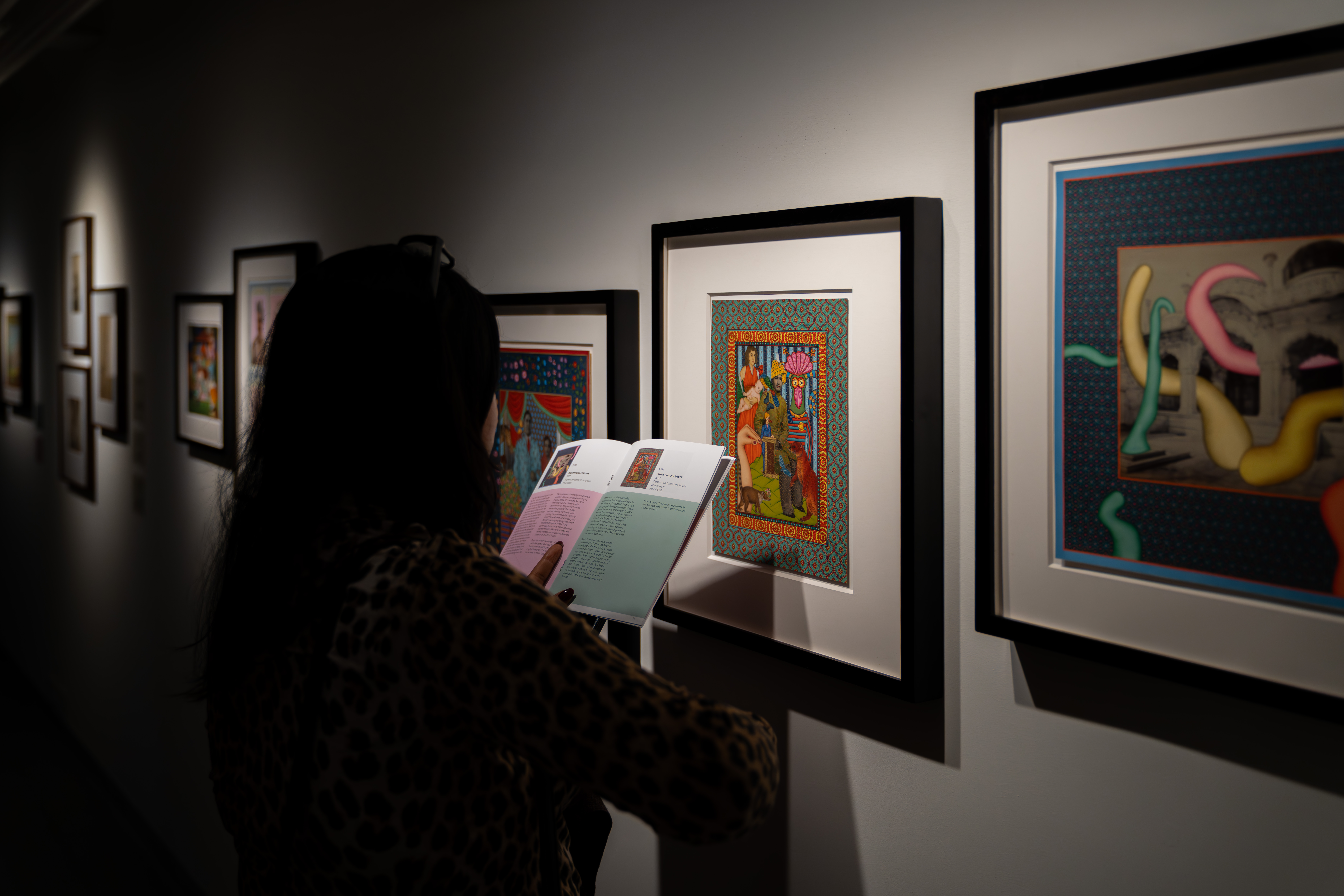 A person looking at an artwork on the wall while reading an exhibition booklet. 