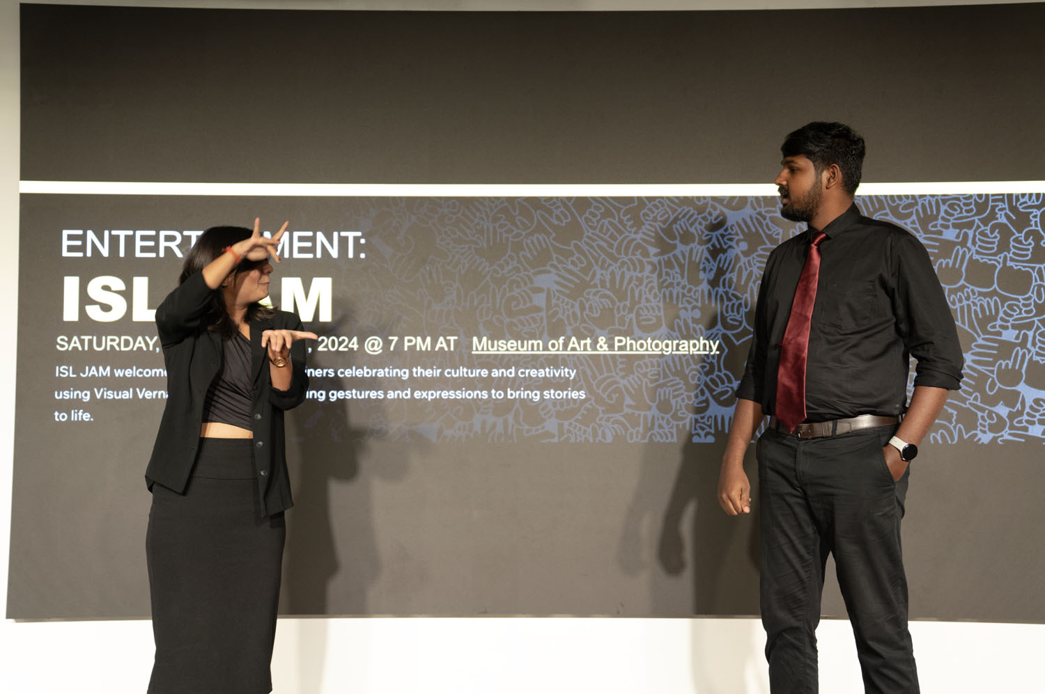 An image of a person dressed in black standing on stage. They are gesturing in ISL.