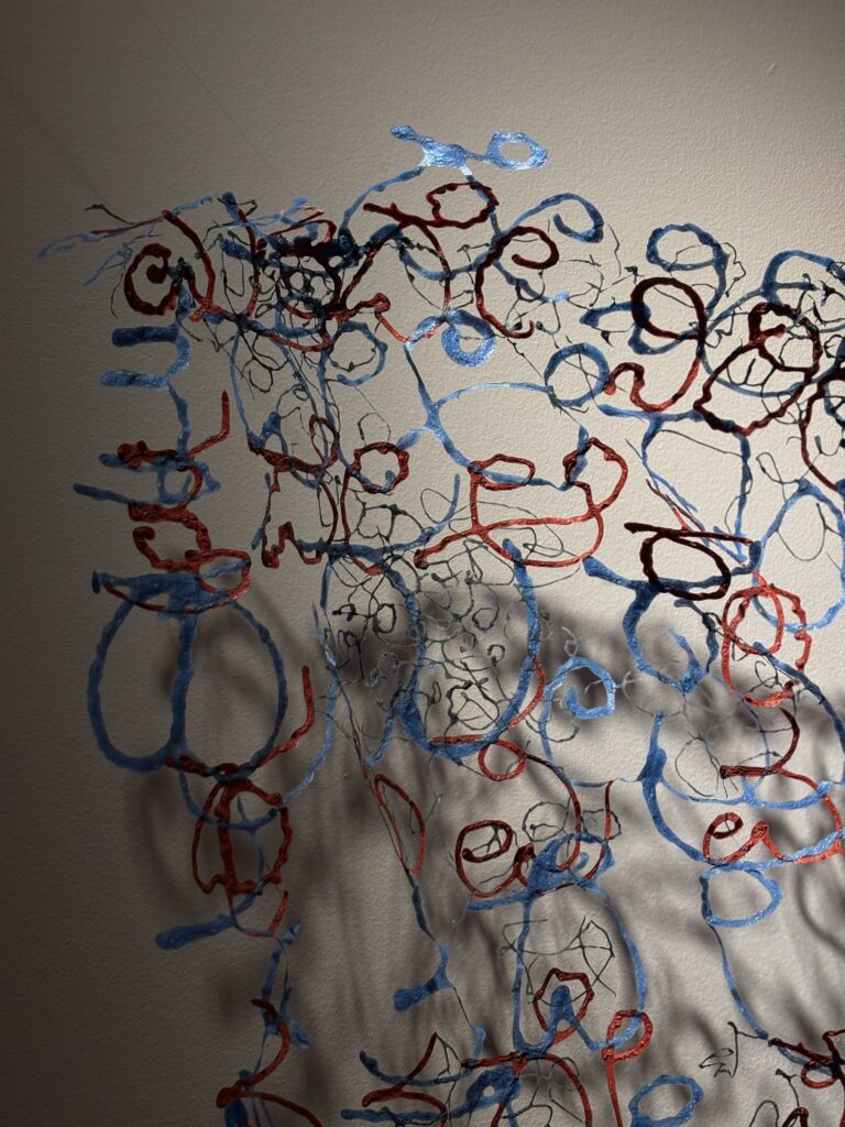 A close-up of an intricate blue and red paper sculpture of repeating swirling patterns and their shadow