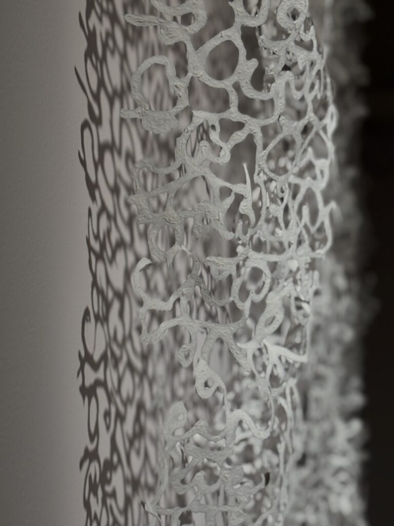 A portion of a white paper sculpture hangs and along with its shadow on the wall, forms a columnar shape 