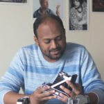 A bust portrait of a man wearing a striped t-shirt while looking at the camera he is holding in his hands 
